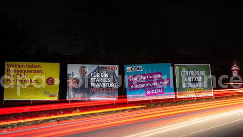 Wahlplakate in Frankfurt am Main neben einer Straße. Es ist Nacht, und die Rücklicher von Autos ziehen sich aufgrund der langen Belichtungszeit als rote Streifen quer über das Foto.