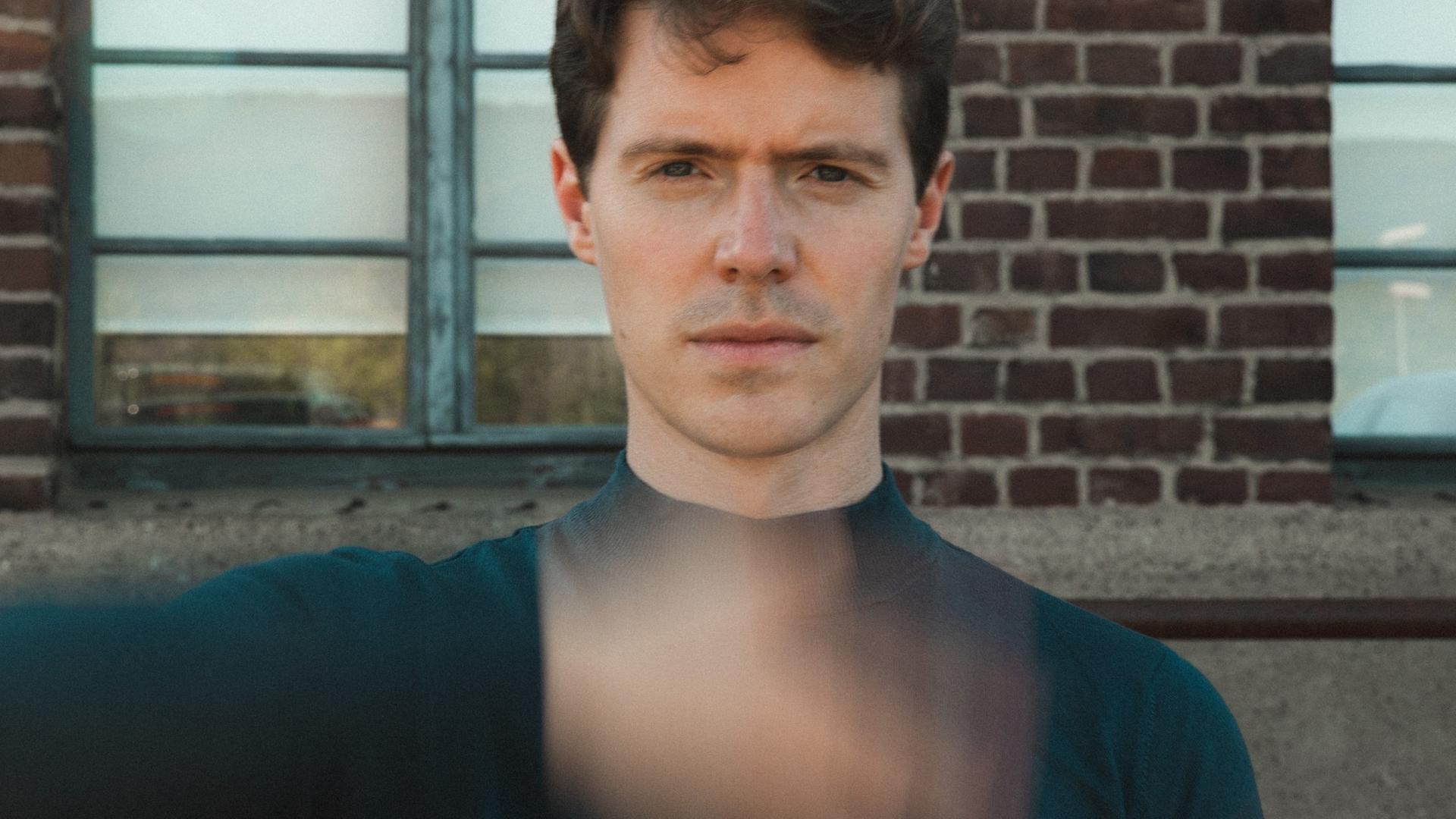 Florian Noack sitzt mit kurzen, dunklen Haaren der Kamera gegenüber und scheint eine Hand direkt vor seinem Körper zu schütteln, die verschwommen vor seinem Oberkörper erscheint. Florian Noack sitzt mit kurzen, dunklen Haaren der Kamera gegenüber und scheint eine Hand direkt vor seinem Körper zu schütteln, die verschwommen vor seinem Oberkörper erscheint.