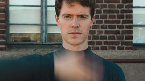 Florian Noack sitzt mit kurzen, dunklen Haaren der Kamera gegenüber und scheint eine Hand direkt vor seinem Körper zu schütteln, die verschwommen vor seinem Oberkörper erscheint.