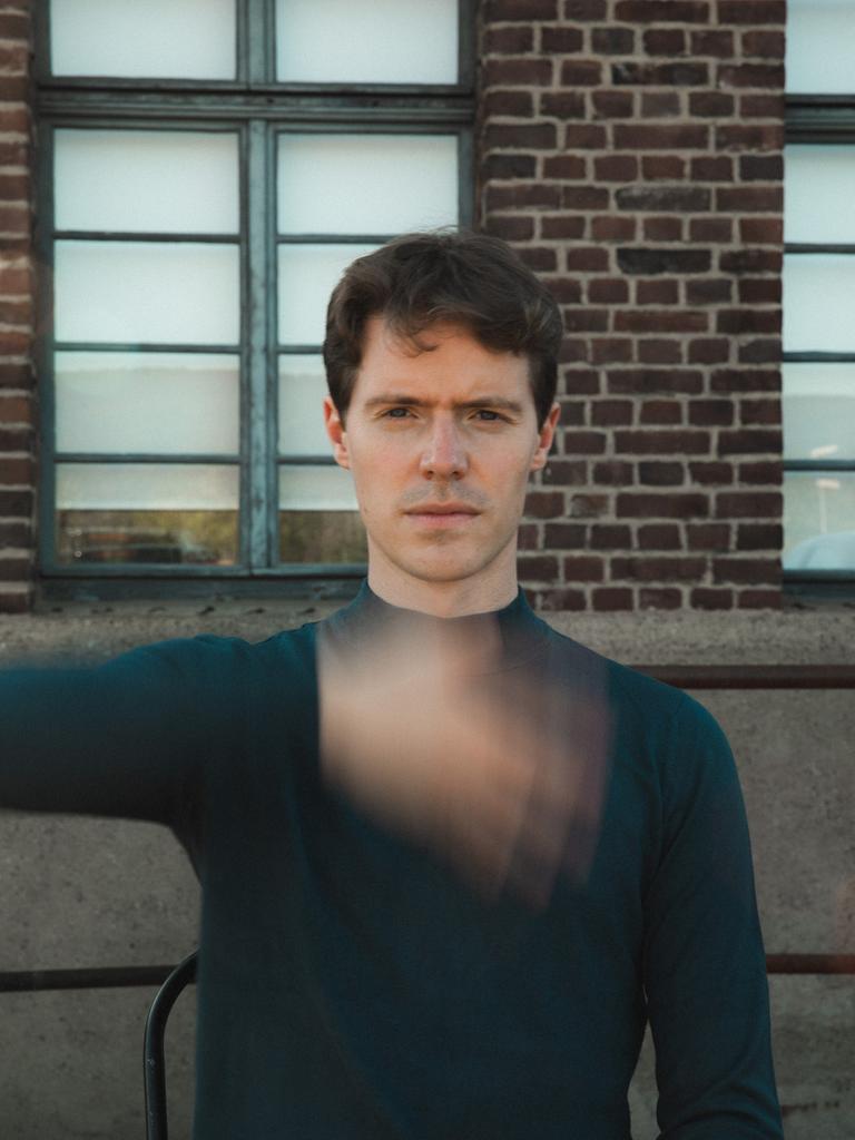 Florian Noack sitzt mit kurzen, dunklen Haaren der Kamera gegenüber und scheint eine Hand direkt vor seinem Körper zu schütteln, die verschwommen vor seinem Oberkörper erscheint.