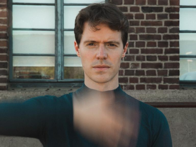 Florian Noack sitzt mit kurzen, dunklen Haaren der Kamera gegenüber und scheint eine Hand direkt vor seinem Körper zu schütteln, die verschwommen vor seinem Oberkörper erscheint.