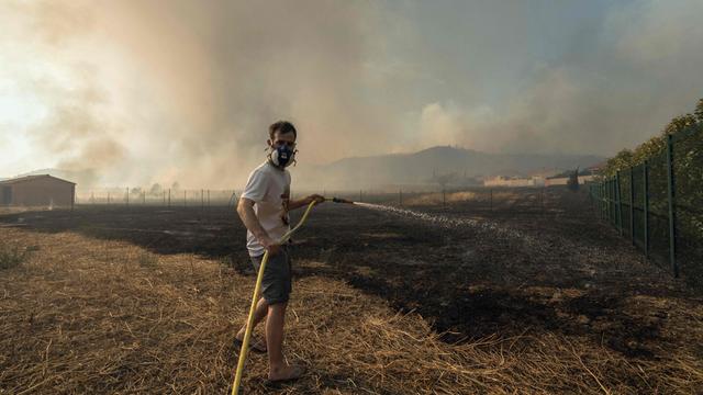 Ein Mann hält einen Schlauch mit Wasser in der Hand. Er trägt eine Schutz-Maske. Vor ihm ist viel verbrannte Erde. In der Luft ist Rauch.