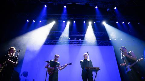 Eine bis zu vierköpfige Band spielt unter blauem Scheinwerferlicht ihre Instrumente, darunter Keyboard und Saxophon.