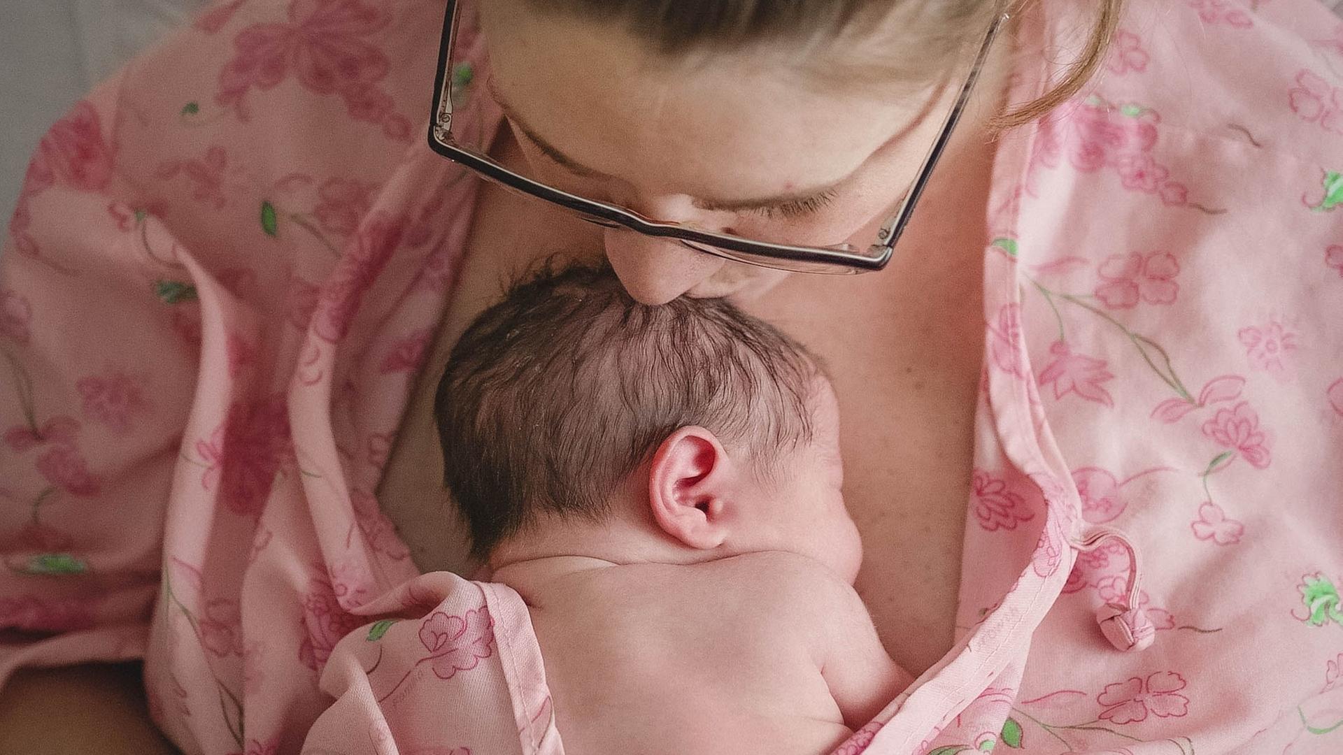 Eine Mutter hat ihr nacktes Baby auf der Brust liegen. Sie trägt einen rosa farbenen Morgenmantel und hat an der Hand Venenzugänge.