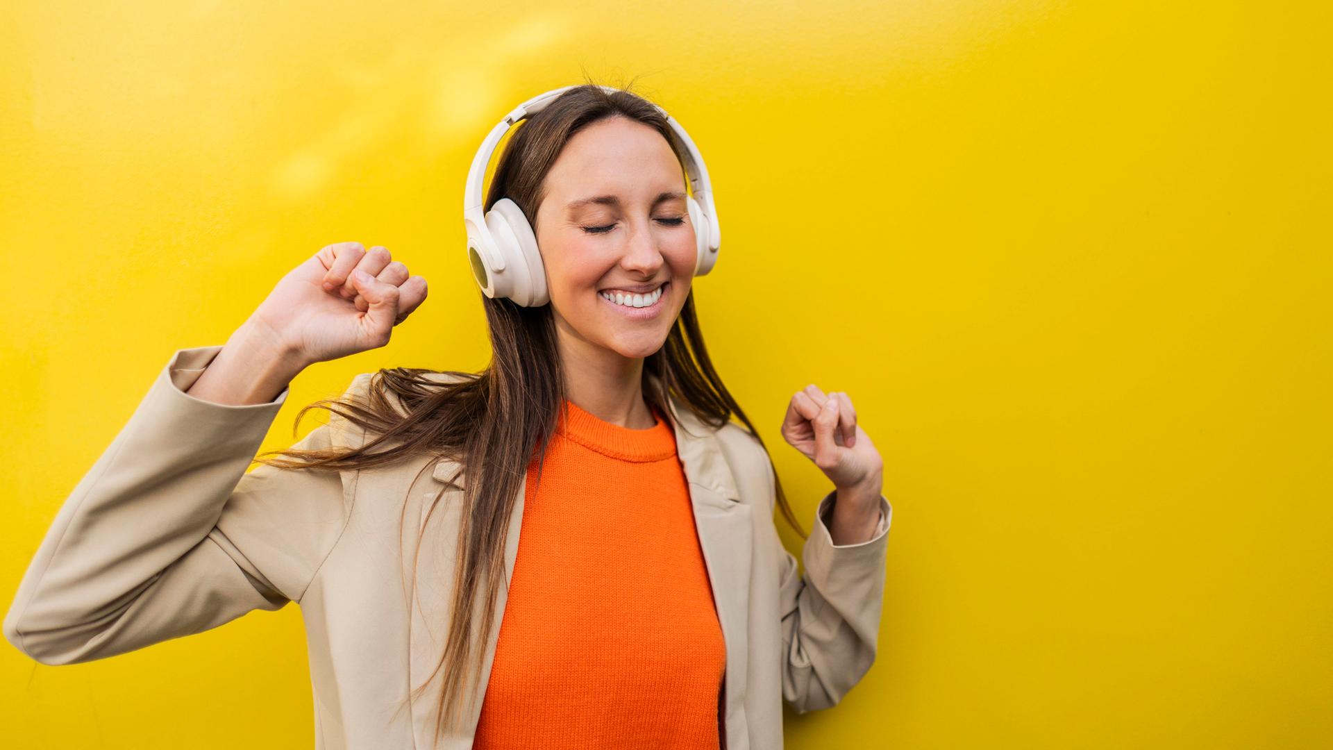 Eine Frau steht tanzend vor einer gelben Wand und tanzt mit geschlossenen Augen