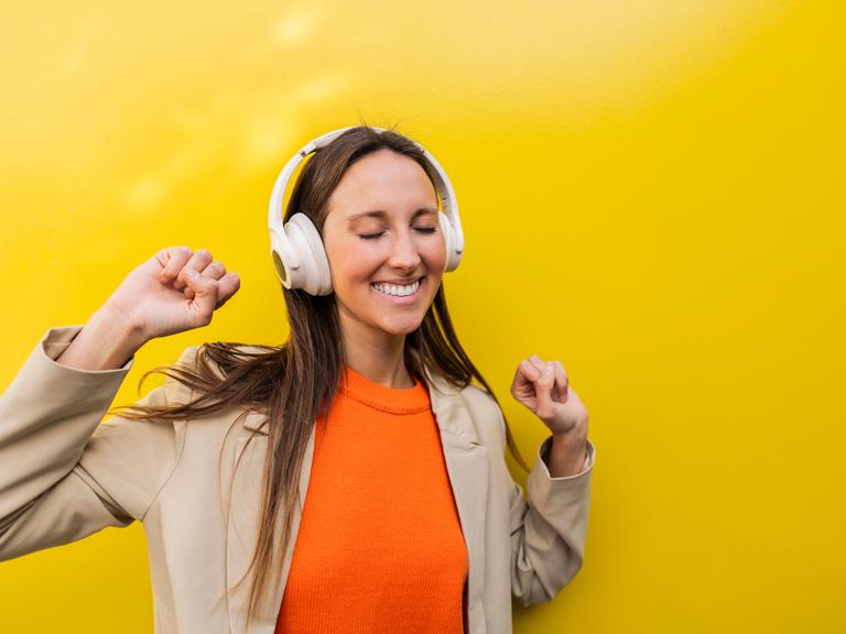 Eine Frau steht tanzend vor einer gelben Wand und tanzt mit geschlossenen Augen