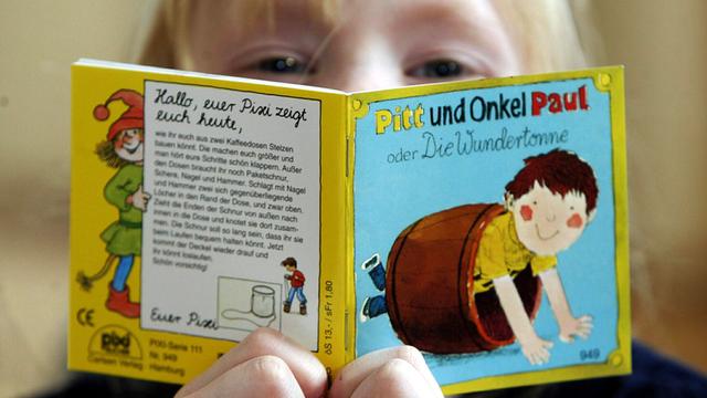Nahaufnahme eines Kindes, das aus einem kleinen, quadratischen Pixi-Buch liest.