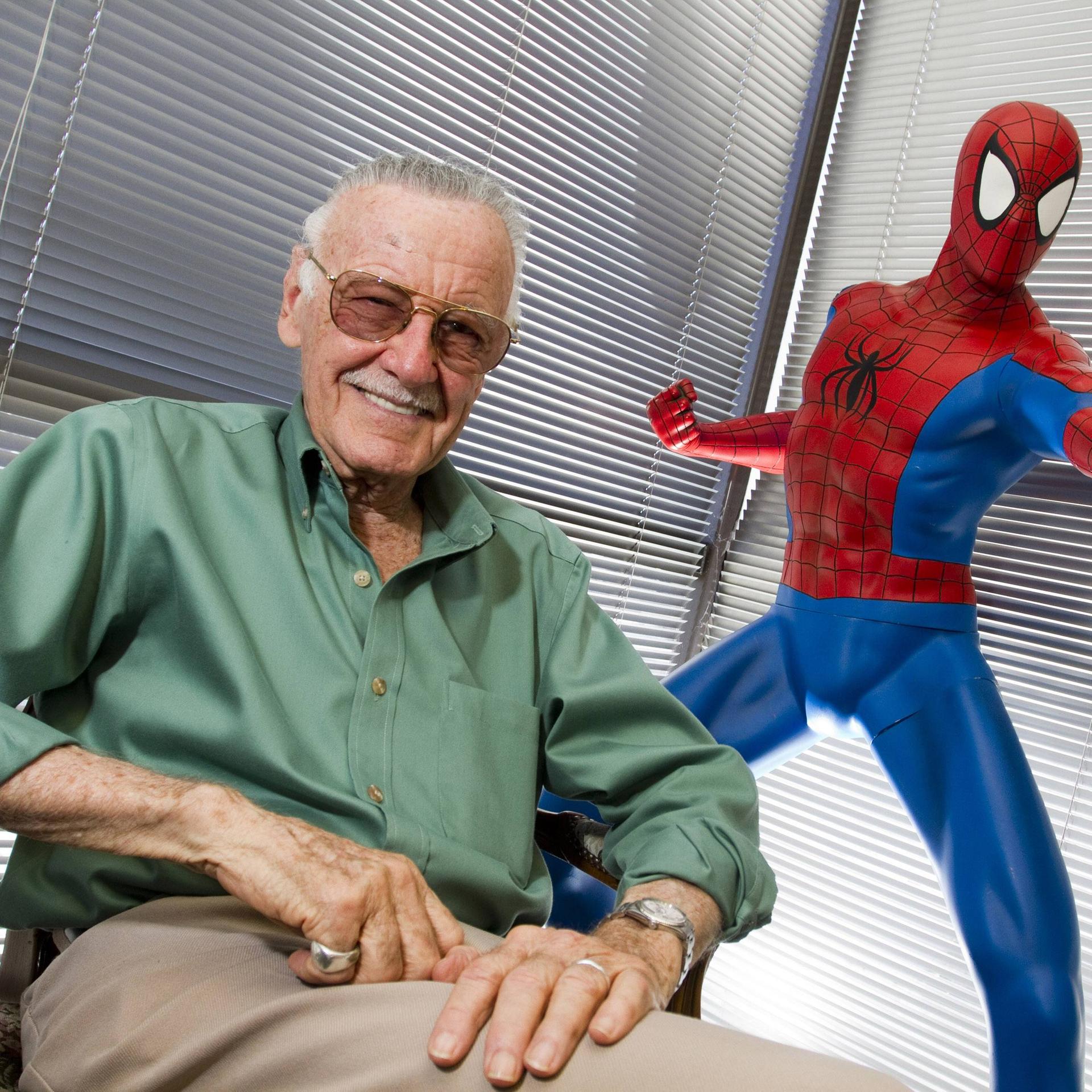 Stan Lee sitzt neben einer Spider-Man-Statue und lacht in die Kamera.