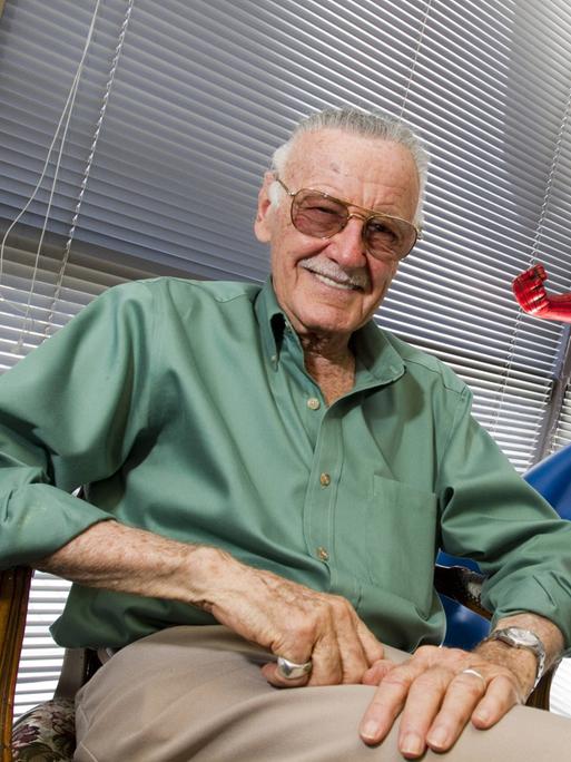 Stan Lee sitzt neben einer Spider-Man-Statue und lacht in die Kamera.
