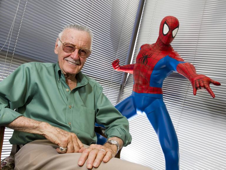 Stan Lee sitzt neben einer Spider-Man-Statue und lacht in die Kamera.