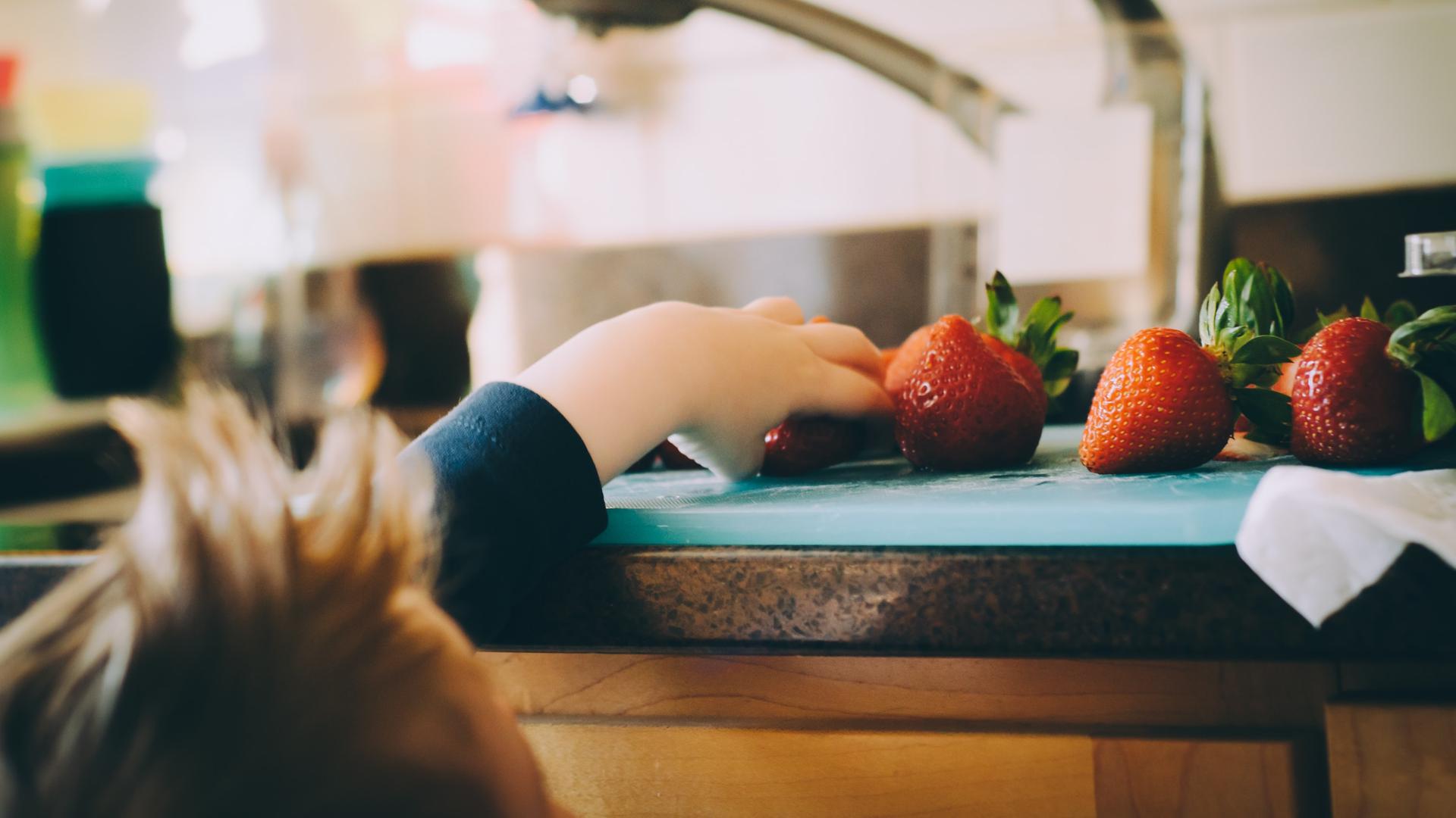 Ein Kind greift von unten nach Erbeeren, die auf einer Küchenarbeitsplatte liegen.