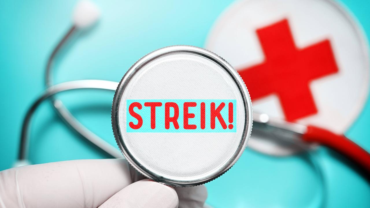 Eine Hand hält ein Stethoskop mit Aufschrift Streik, Symbolfoto Ärztestreik. Im Hintergrund ist ein Rotes Kreuz zu sehen.