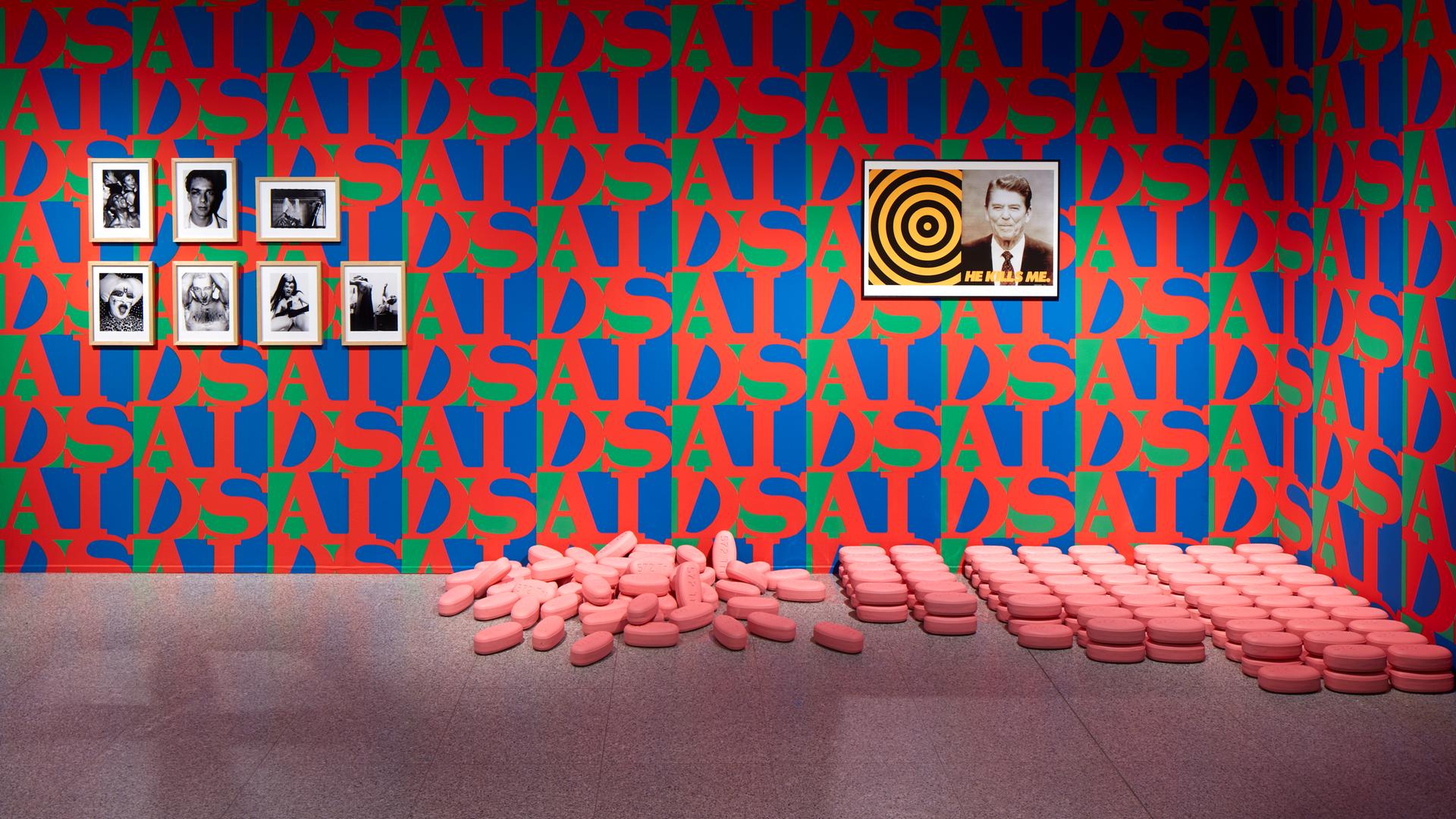 Die Wände eines Ausstellungsraumes sind mit rot-blau-grüner Tapete tapeziert. Der Schriftzug AIDS wiederholt sich und bildet ein Muster. An der Wand hängt ein Bild von J. F. Kennedy, am Boden liegen überdimensional große rosa Pillen.