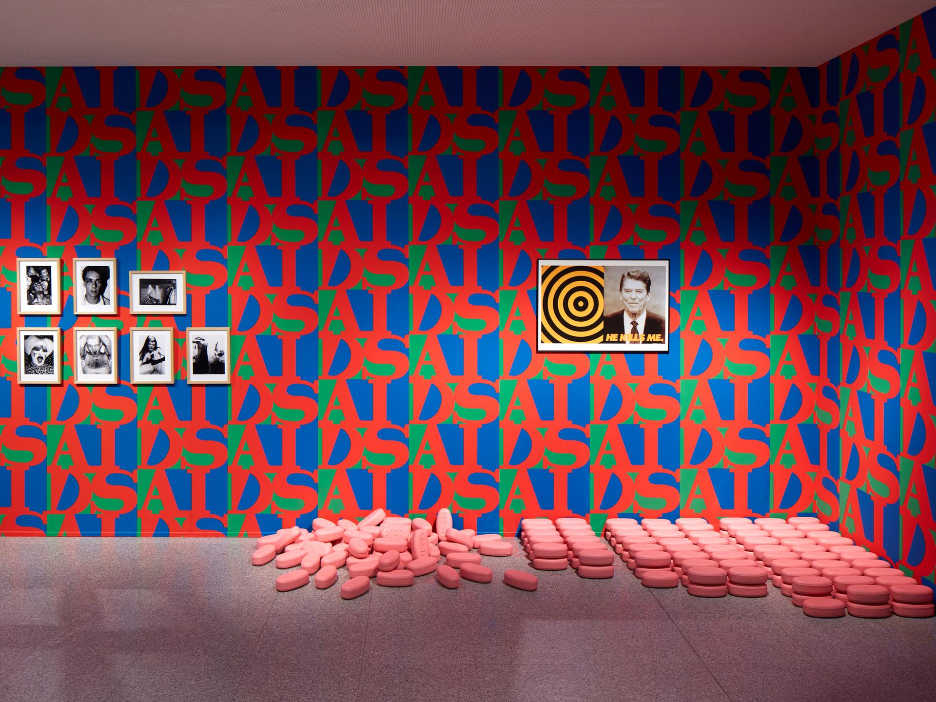 Die Wände eines Ausstellungsraumes sind mit rot-blau-grüner Tapete tapeziert. Der Schriftzug AIDS wiederholt sich und bildet ein Muster. An der Wand hängt ein Bild von J. F. Kennedy, am Boden liegen überdimensional große rosa Pillen.