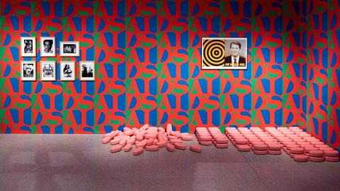 Die Wände eines Ausstellungsraumes sind mit rot-blau-grüner Tapete tapeziert. Der Schriftzug AIDS wiederholt sich und bildet ein Muster. An der Wand hängt ein Bild von J. F. Kennedy, am Boden liegen überdimensional große rosa Pillen.