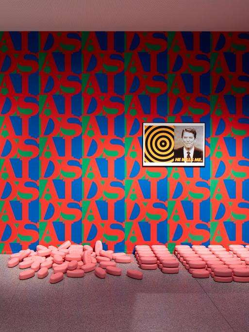 Die Wände eines Ausstellungsraumes sind mit rot-blau-grüner Tapete tapeziert. Der Schriftzug AIDS wiederholt sich und bildet ein Muster. An der Wand hängt ein Bild von J. F. Kennedy, am Boden liegen überdimensional große rosa Pillen.