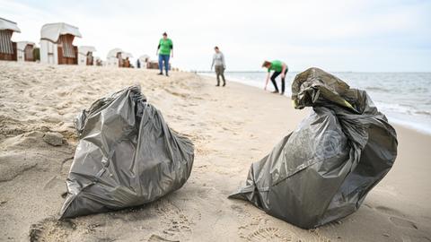 Müllsäcke stehen an einem Strand, von dem im Hintergrund drei Jugendliche Müll aufsammeln.