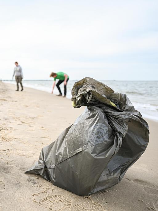 Müllsäcke stehen an einem Strand, von dem im Hintergrund drei Jugendliche Müll aufsammeln.