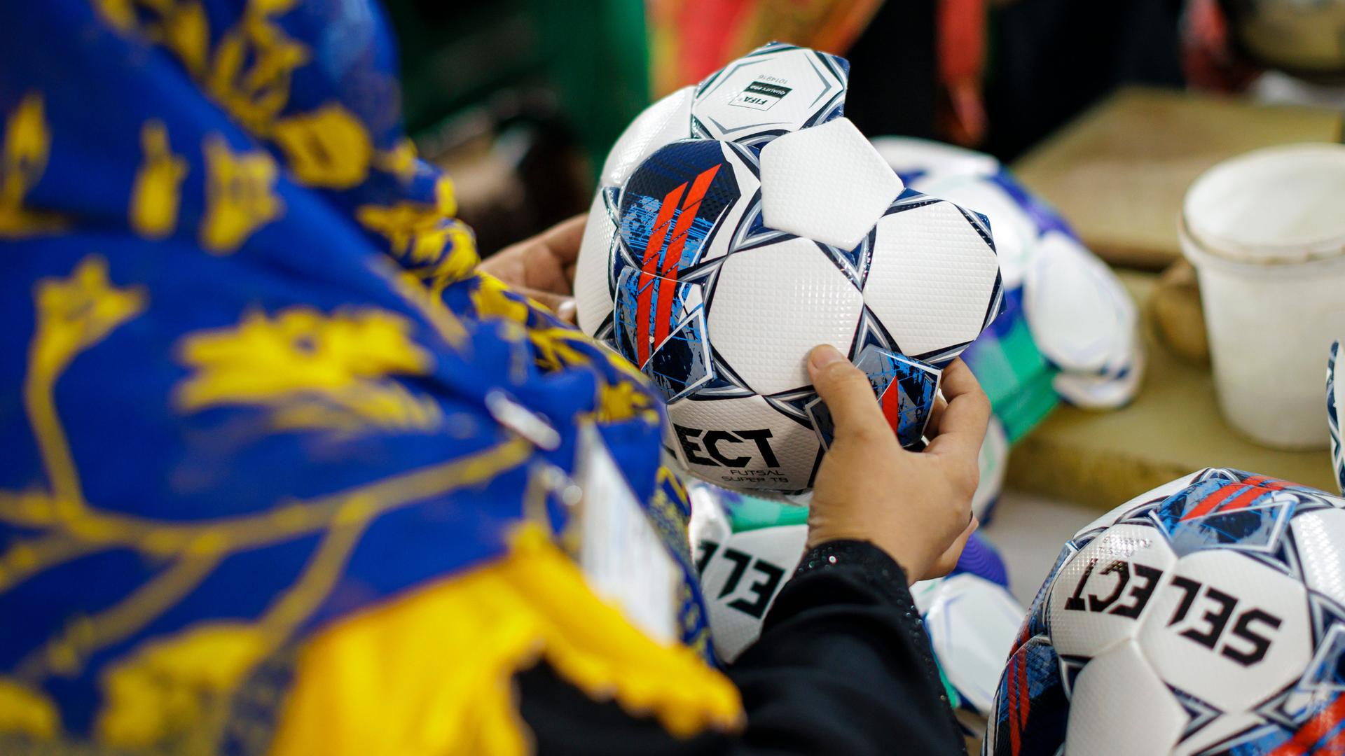 Aufnahme einer Person, die einen noch nicht fertiggestellten Fußball in der Hand hält. Die Person trägt ein farbenfrohes Kopftuch und wird von hinten gezeigt. Somit ist ihr Gesicht nicht zu erkennen. Aufnahme einer Person, die einen noch nicht fertiggestellten Fußball in der Hand hält. Die Person trägt ein farbenfrohes Kopftuch und wird von hinten gezeigt. Somit ist ihr Gesicht nicht zu erkennen.