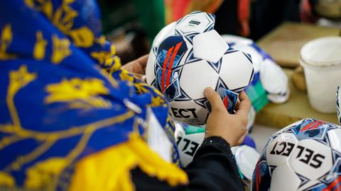 Aufnahme einer Person, die einen noch nicht fertiggestellten Fußball in der Hand hält. Die Person trägt ein farbenfrohes Kopftuch und wird von hinten gezeigt. Somit ist ihr Gesicht nicht zu erkennen. Aufnahme einer Person, die einen noch nicht fertiggestellten Fußball in der Hand hält. Die Person trägt ein farbenfrohes Kopftuch und wird von hinten gezeigt. Somit ist ihr Gesicht nicht zu erkennen.