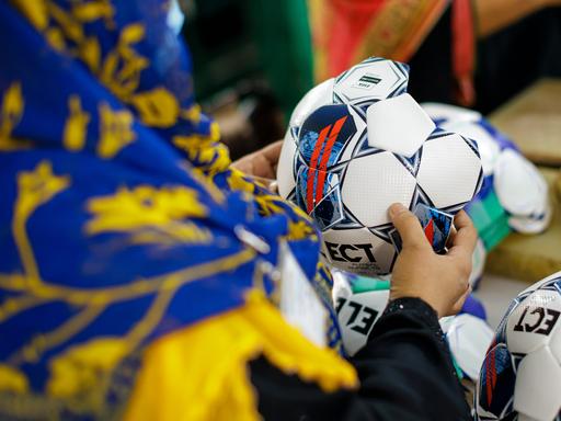 Aufnahme einer Person, die einen noch nicht fertiggestellten Fußball in der Hand hält. Die Person trägt ein farbenfrohes Kopftuch und wird von hinten gezeigt. Somit ist ihr Gesicht nicht zu erkennen.