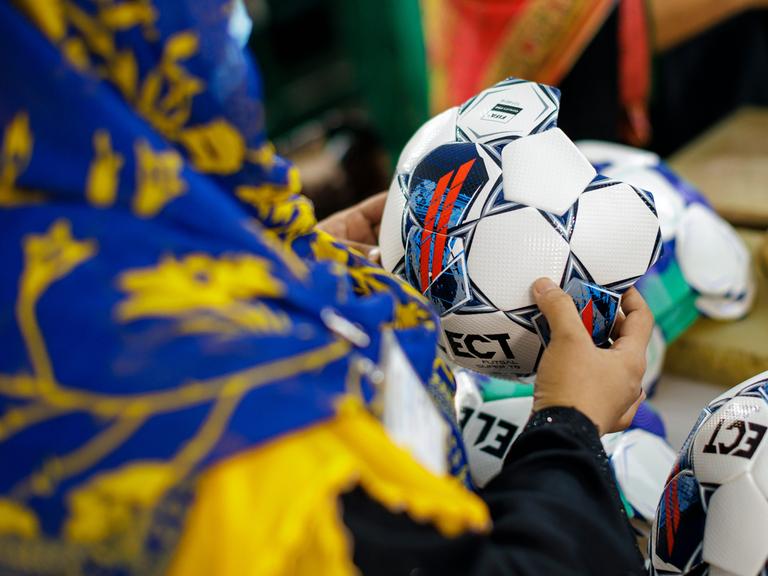 Aufnahme einer Person, die einen noch nicht fertiggestellten Fußball in der Hand hält. Die Person trägt ein farbenfrohes Kopftuch und wird von hinten gezeigt. Somit ist ihr Gesicht nicht zu erkennen.