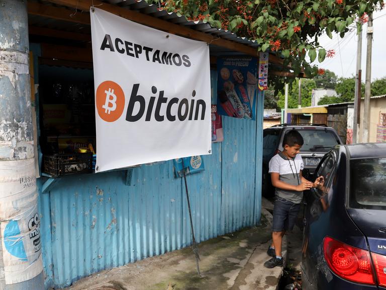 Ein Jugendlicher steht neben einem Auto, während er vor einem kleinen Geschäft wartet, das Bitcoin akzeptiert.