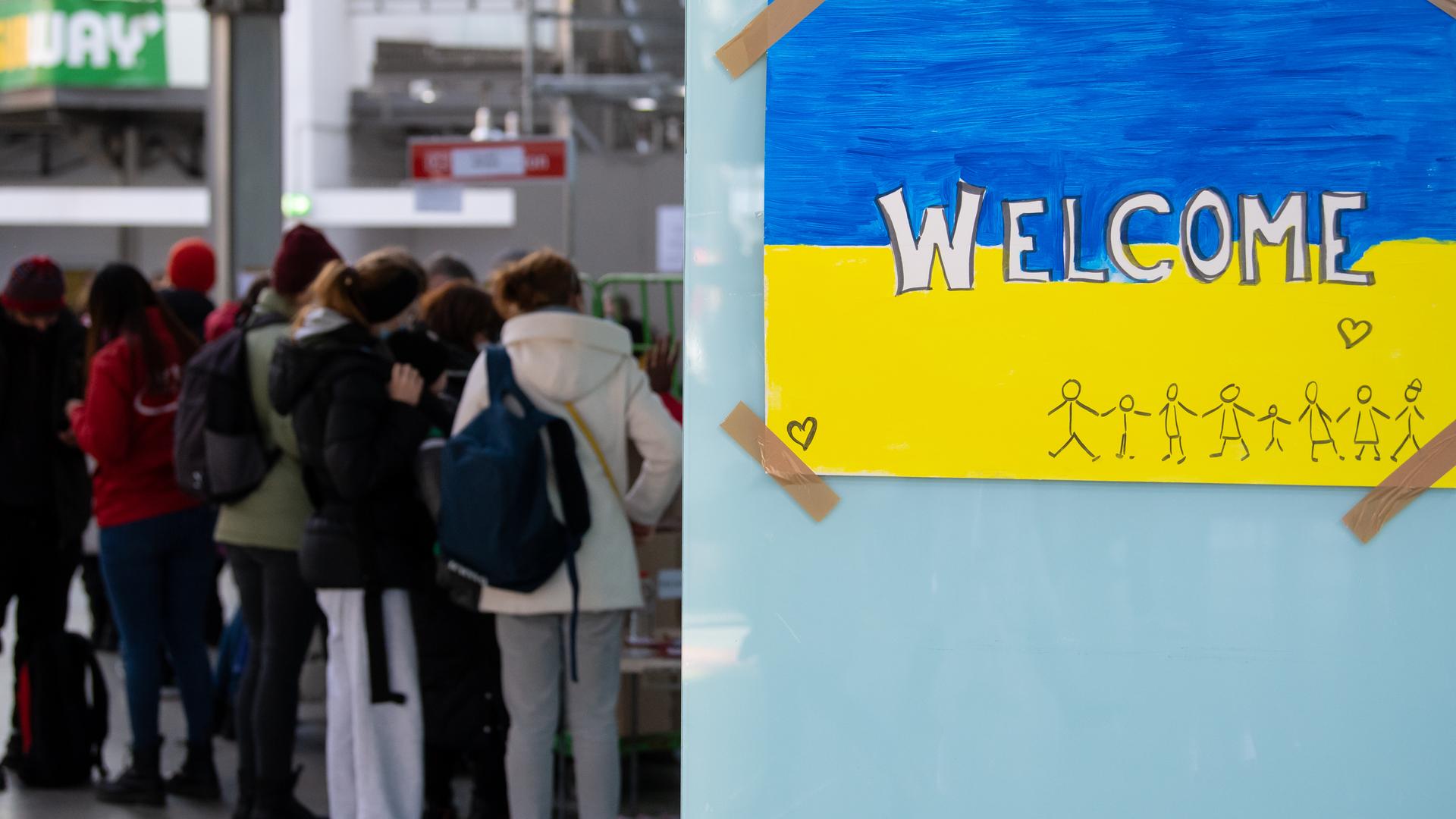 Ein Schild in den Farben der Ukraine mit der Aufschrift "Welcome" ist am Hauptbahnhof zu sehen. Im Hintergrund werden Geflüchtete aus der Ukraine nach ihrer Ankunft von Mitarbeitenden der Caritas und Ehrenamtlichen empfangen.