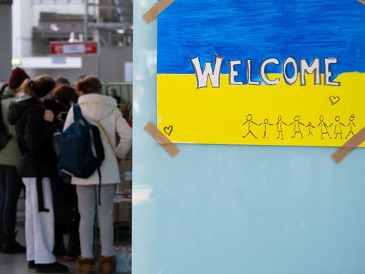 Ein Schild in den Farben der Ukraine mit der Aufschrift "Welcome" ist am Hauptbahnhof zu sehen. Im Hintergrund werden Geflüchtete aus der Ukraine nach ihrer Ankunft von Mitarbeitenden der Caritas und Ehrenamtlichen empfangen.