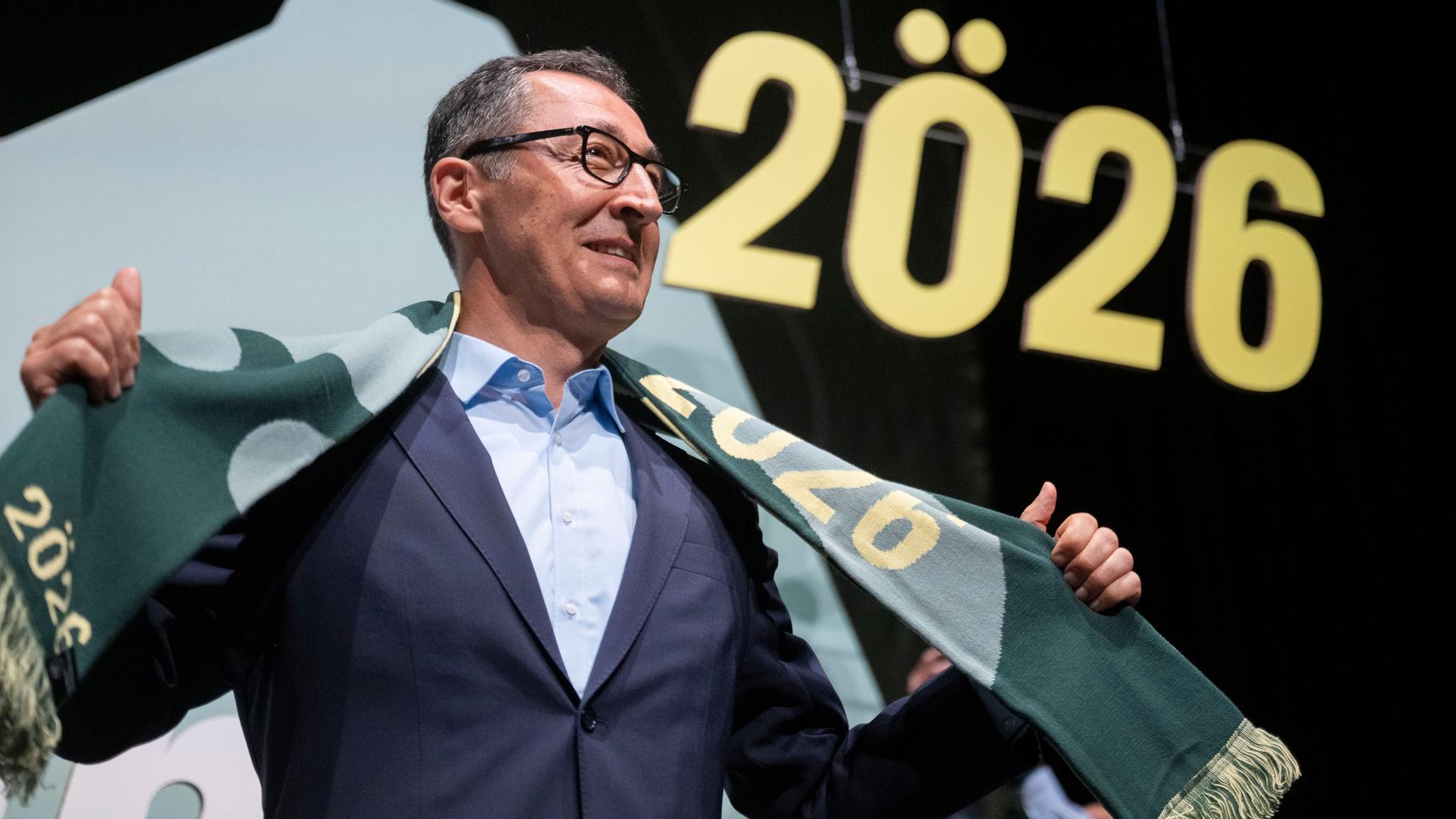 Der Grünen-Politiker Cem Özdemir steht lächelnd auf einer Bühne und hält einen Schal in den Händen.