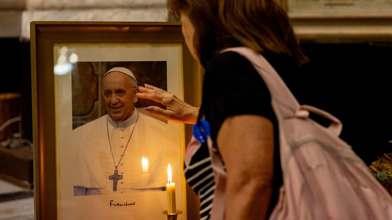 Ein Foto von Papst Franziskus steht mit einer Kerze davor in einer KIrche und eine Frau steht davor. 