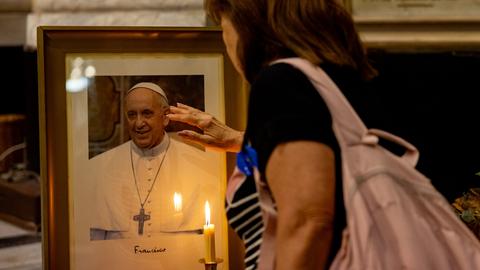 Ein Foto von Papst Franziskus steht mit einer Kerze davor in einer KIrche und eine Frau steht davor. 
