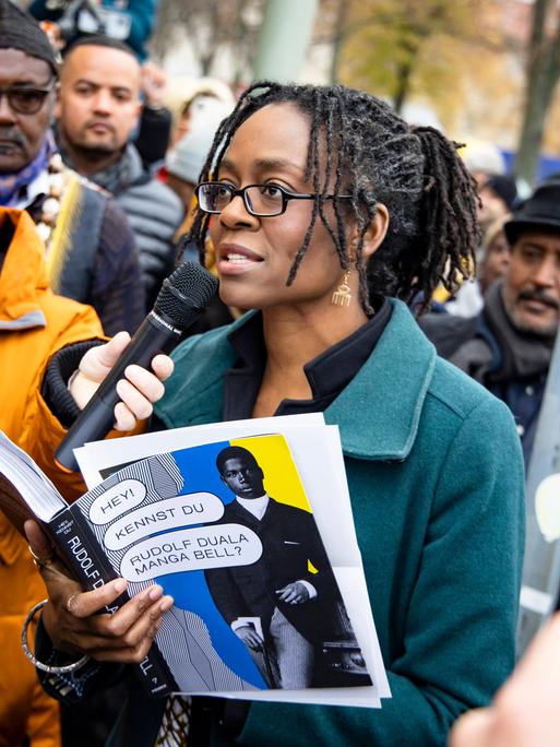 Sharon Dodua Otoo steht auf einer Straßenkreuzung und spricht einen Text in ein Mikrofon. Um sie herum steht eine Menschenmenge.