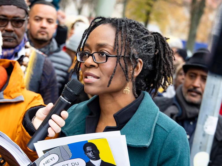 Sharon Dodua Otoo steht auf einer Straßenkreuzung und spricht einen Text in ein Mikrofon. Um sie herum steht eine Menschenmenge.