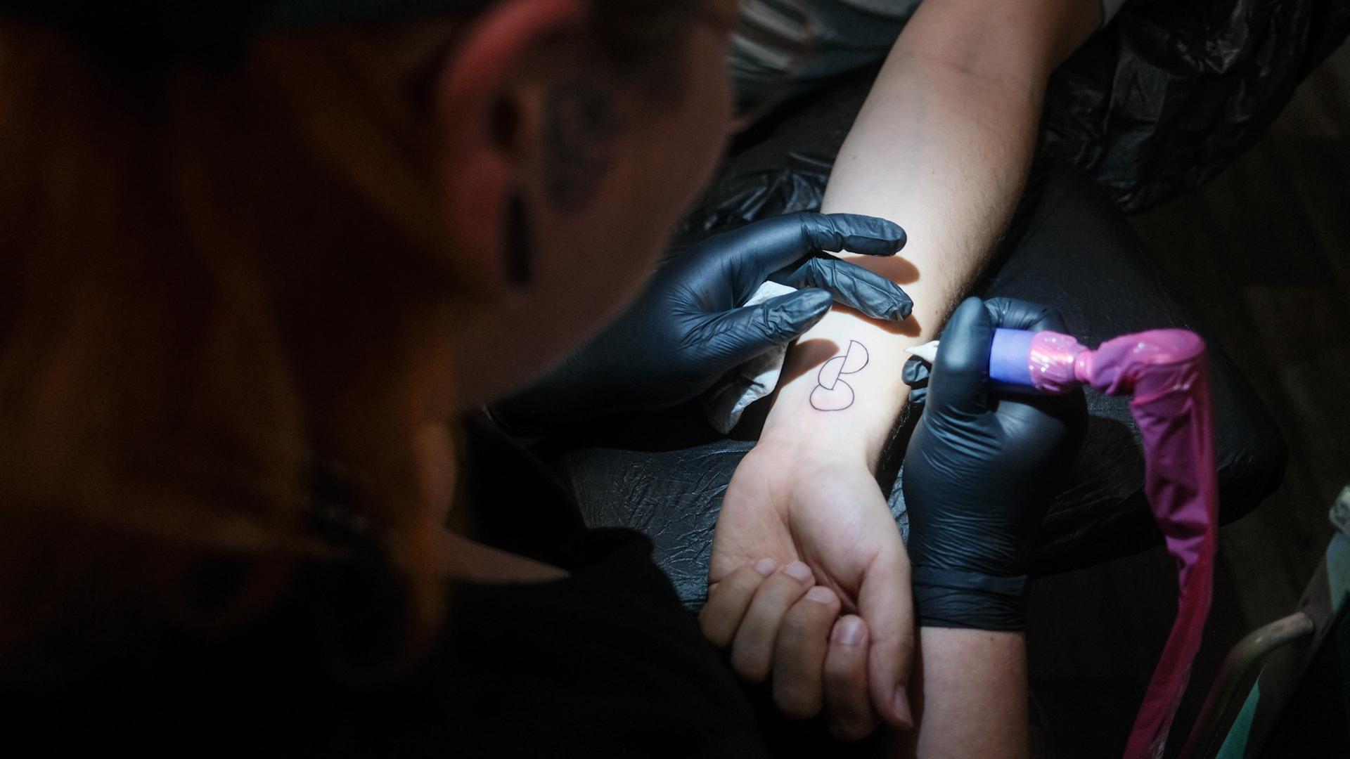 Eine Tätowiererin sticht das OrganspendeTattoo auf einen Unterarm. Es zeigt drei übereinander verschobene Kreise und Halbkreise. Eine Tätowiererin sticht das OrganspendeTattoo auf einen Unterarm. Es zeigt drei übereinander verschobene Kreise und Halbkreise.