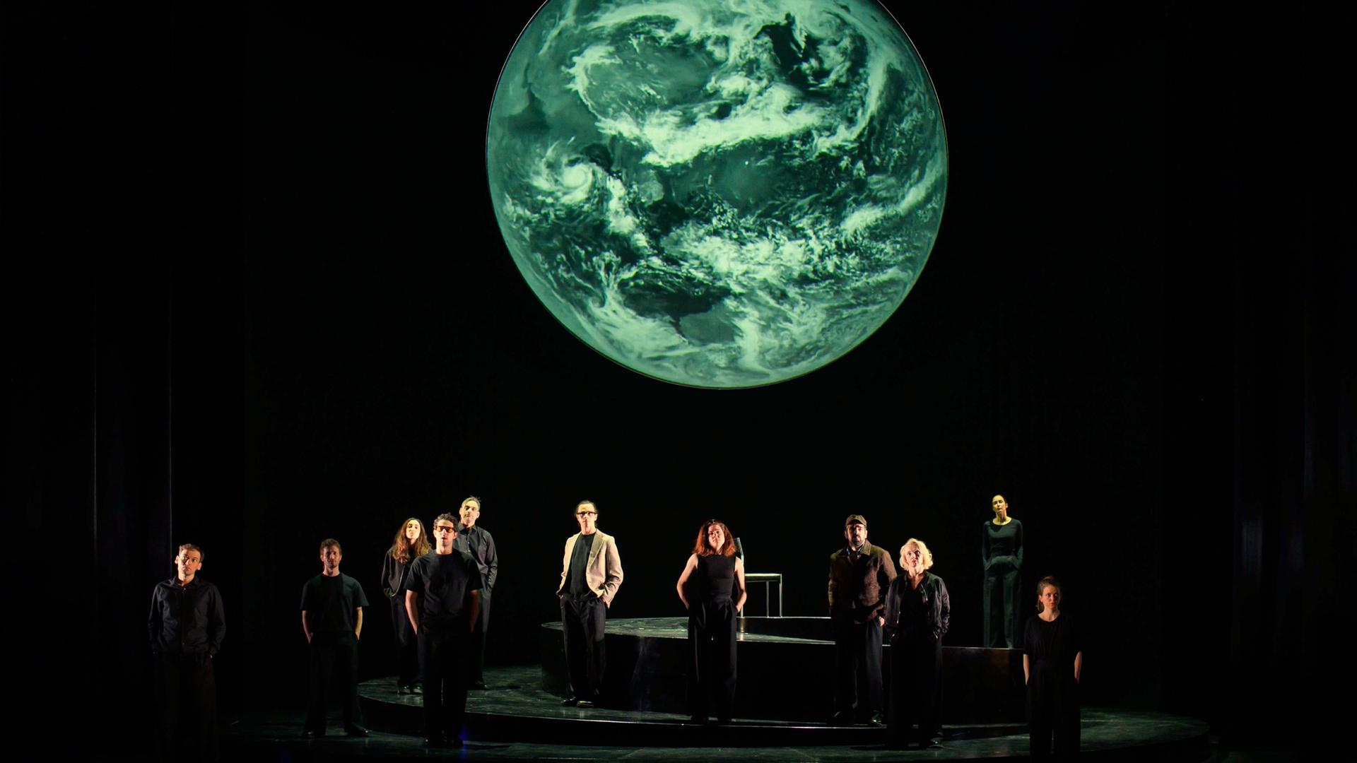 Szenenfoto aus der Produktion "Wir sind das Volk" am Deutschen Nationaltheater Weimar: Schauspielende auf einer dunklen Bühne blicken Richtung Publikum, hinter ihnen eine Projektion der Erdkugel. Szenenfoto aus der Produktion "Wir sind das Volk" am Deutschen Nationaltheater Weimar: Schauspielende auf einer dunklen Bühne blicken Richtung Publikum, hinter ihnen eine Projektion der Erdkugel.