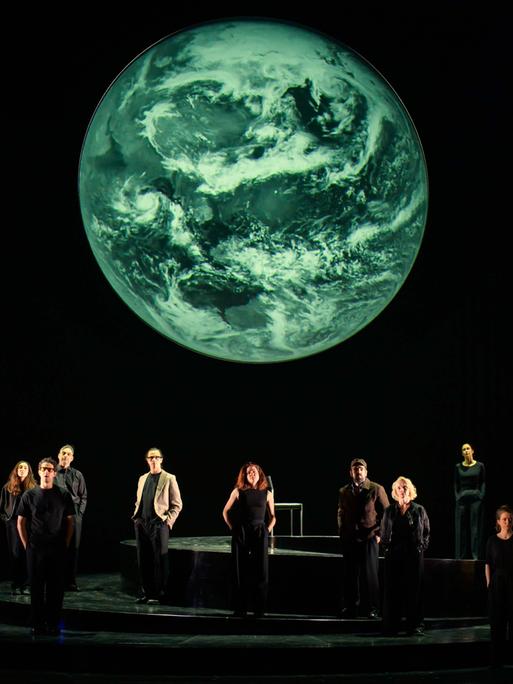 Szenenfoto aus der Produktion "Wir sind das Volk" am Deutschen Nationaltheater Weimar: Schauspielende auf einer dunklen Bühne blicken Richtung Publikum, hinter ihnen eine Projektion der Erdkugel. Szenenfoto aus der Produktion "Wir sind das Volk" am Deutschen Nationaltheater Weimar: Schauspielende auf einer dunklen Bühne blicken Richtung Publikum, hinter ihnen eine Projektion der Erdkugel.