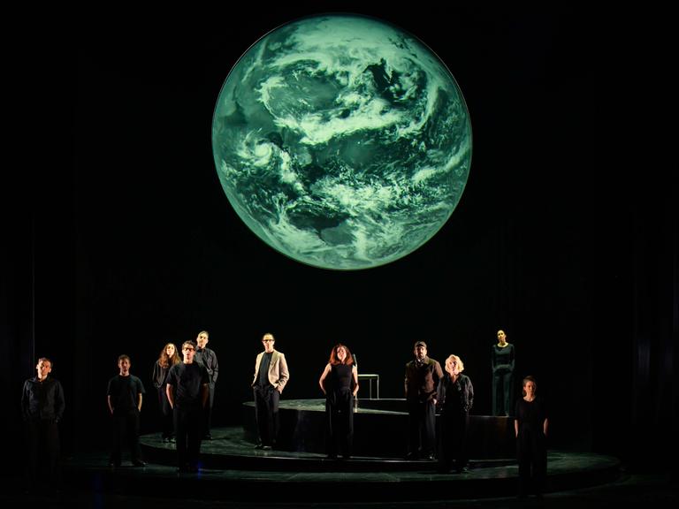 Szenenfoto aus der Produktion "Wir sind das Volk" am Deutschen Nationaltheater Weimar: Schauspielende auf einer dunklen Bühne blicken Richtung Publikum, hinter ihnen eine Projektion der Erdkugel. Szenenfoto aus der Produktion "Wir sind das Volk" am Deutschen Nationaltheater Weimar: Schauspielende auf einer dunklen Bühne blicken Richtung Publikum, hinter ihnen eine Projektion der Erdkugel.