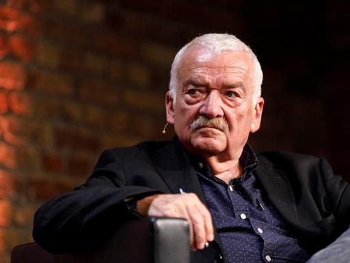 Der Kulturwissenschaftler Hans Ulrich Gumbrecht sitzt in einem schwarzen Sessel mit hochgezogemem Knie. 