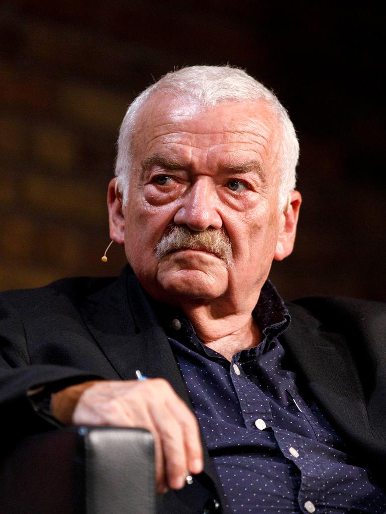 Der Kulturwissenschaftler Hans Ulrich Gumbrecht sitzt in einem schwarzen Sessel mit hochgezogemem Knie. 