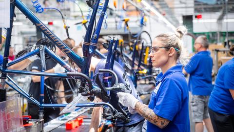Eine blonde Frau mit blauem T-Shirt trägt eine Schutzbrille und baut in einer Werkshalle ein neues Fahrrad zusammen.