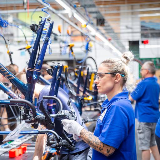 Eine blonde Frau mit blauem T-Shirt trägt eine Schutzbrille und baut in einer Werkshalle ein neues Fahrrad zusammen.