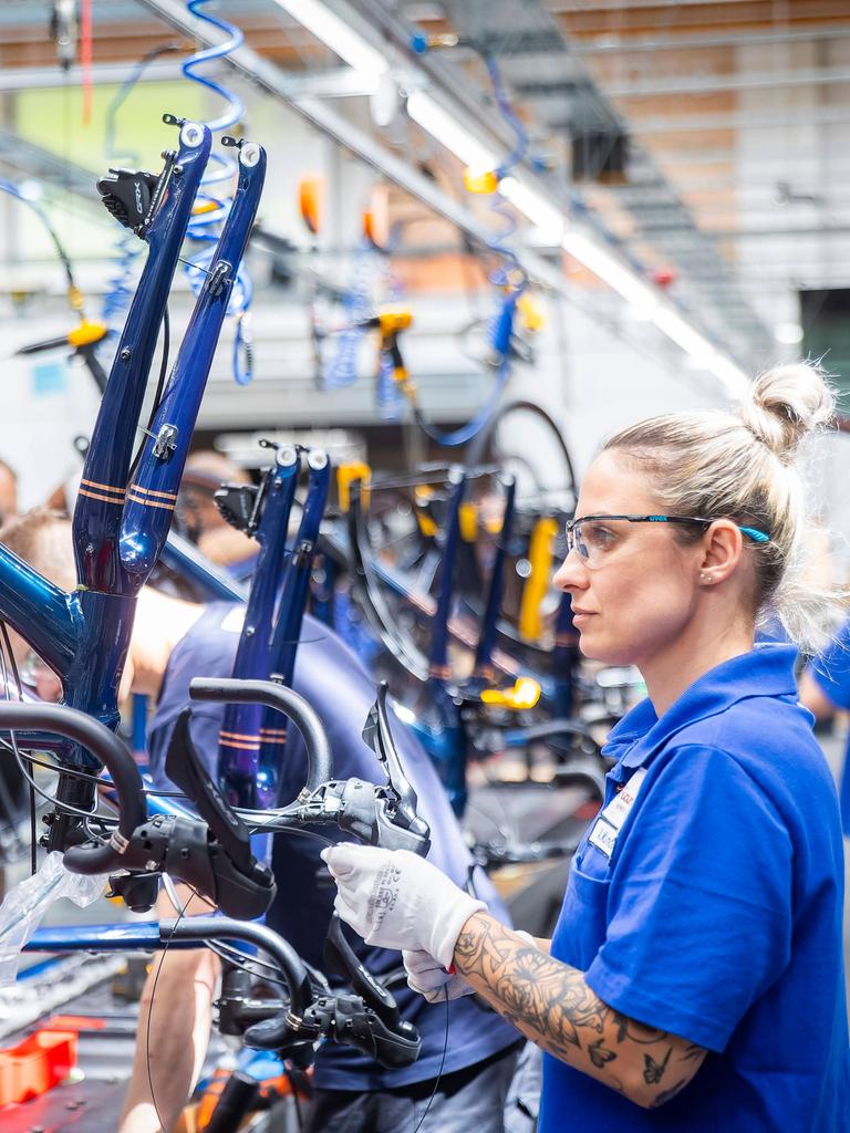Eine blonde Frau mit blauem T-Shirt trägt eine Schutzbrille und baut in einer Werkshalle ein neues Fahrrad zusammen.