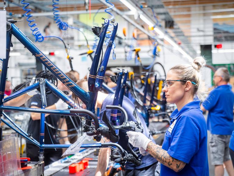 Eine blonde Frau mit blauem T-Shirt trägt eine Schutzbrille und baut in einer Werkshalle ein neues Fahrrad zusammen.