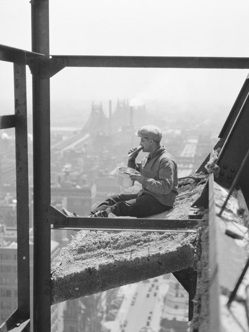 Eine historische Schwarz-Weiß-Aufnahme aus dem Jahr 1922. Ein Maler sitzt zur Mittagszeit auf einem Sims etwa 30 Stockwerke über dem Niveau der Seitenwände eines im Bau befindlichen Gebäudes.