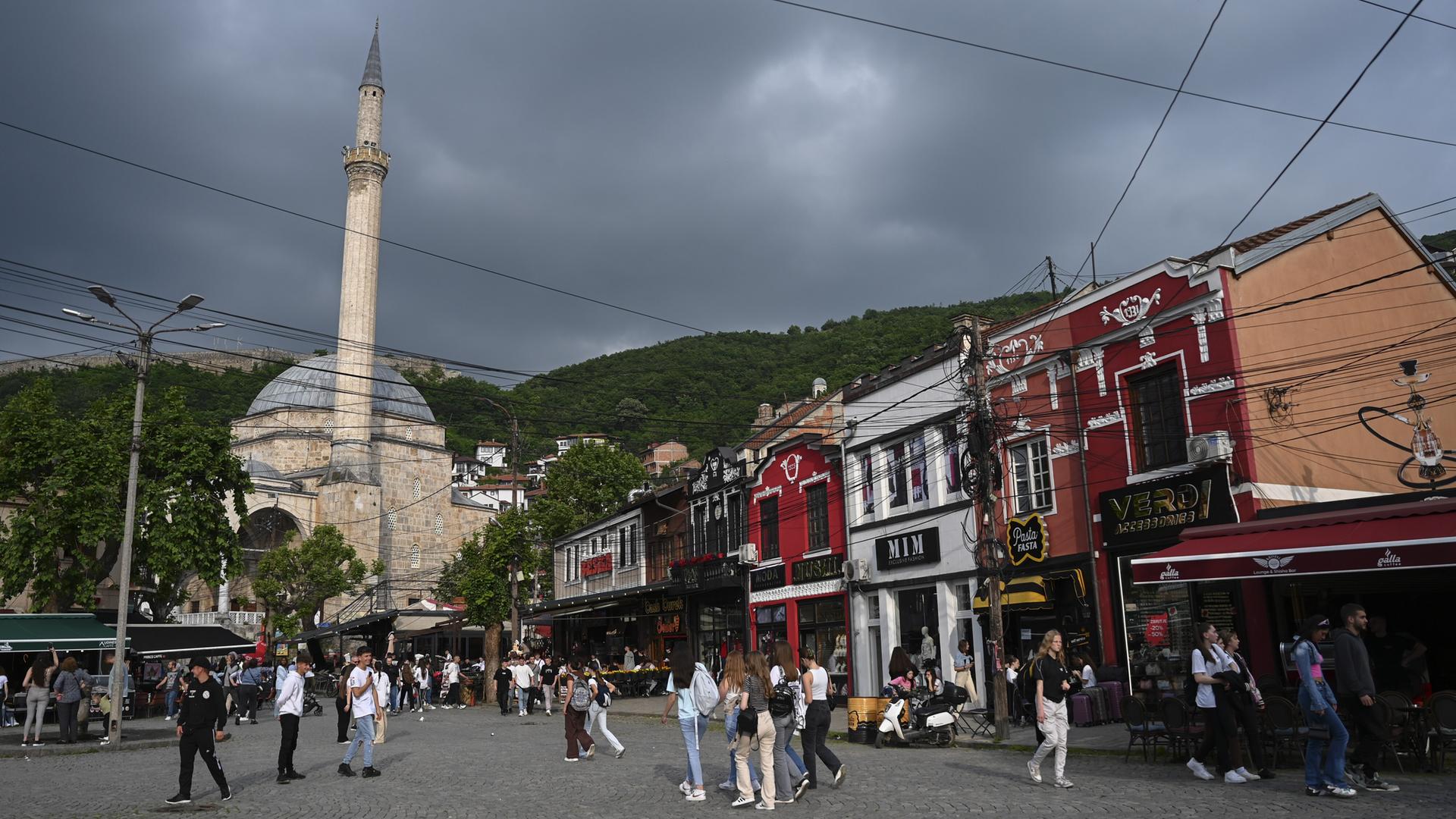 Hinter einem Platz mit Menschen erhebt sich eine weiße Moschee mit weißem Turm. Im Hintergrund: grüne Hügel und ein dunkler Himmel. Hinter einem Platz mit Menschen erhebt sich eine weiße Moschee mit weißem Turm. Im Hintergrund: grüne Hügel und ein dunkler Himmel.