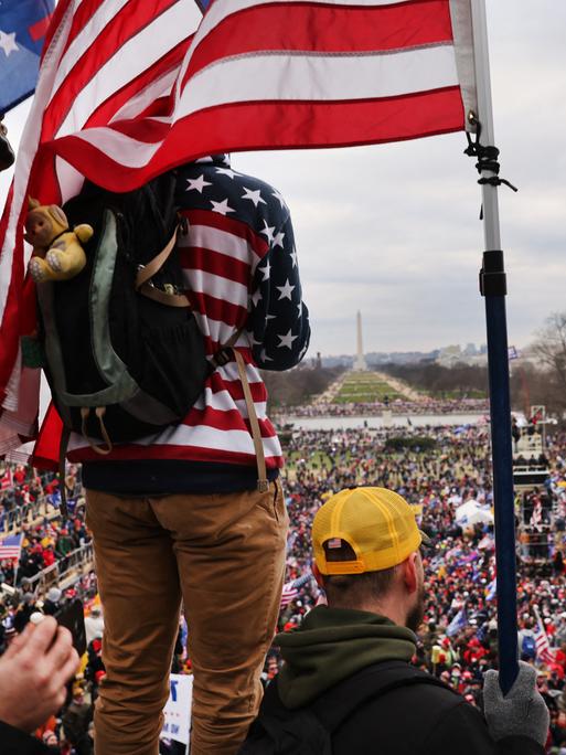Demonstranten mit US-Flaggen stehen auf der Terrasse des Kapitols und überblicken den Vorplatz, auf dem sich eine Menschenmenge drängt.