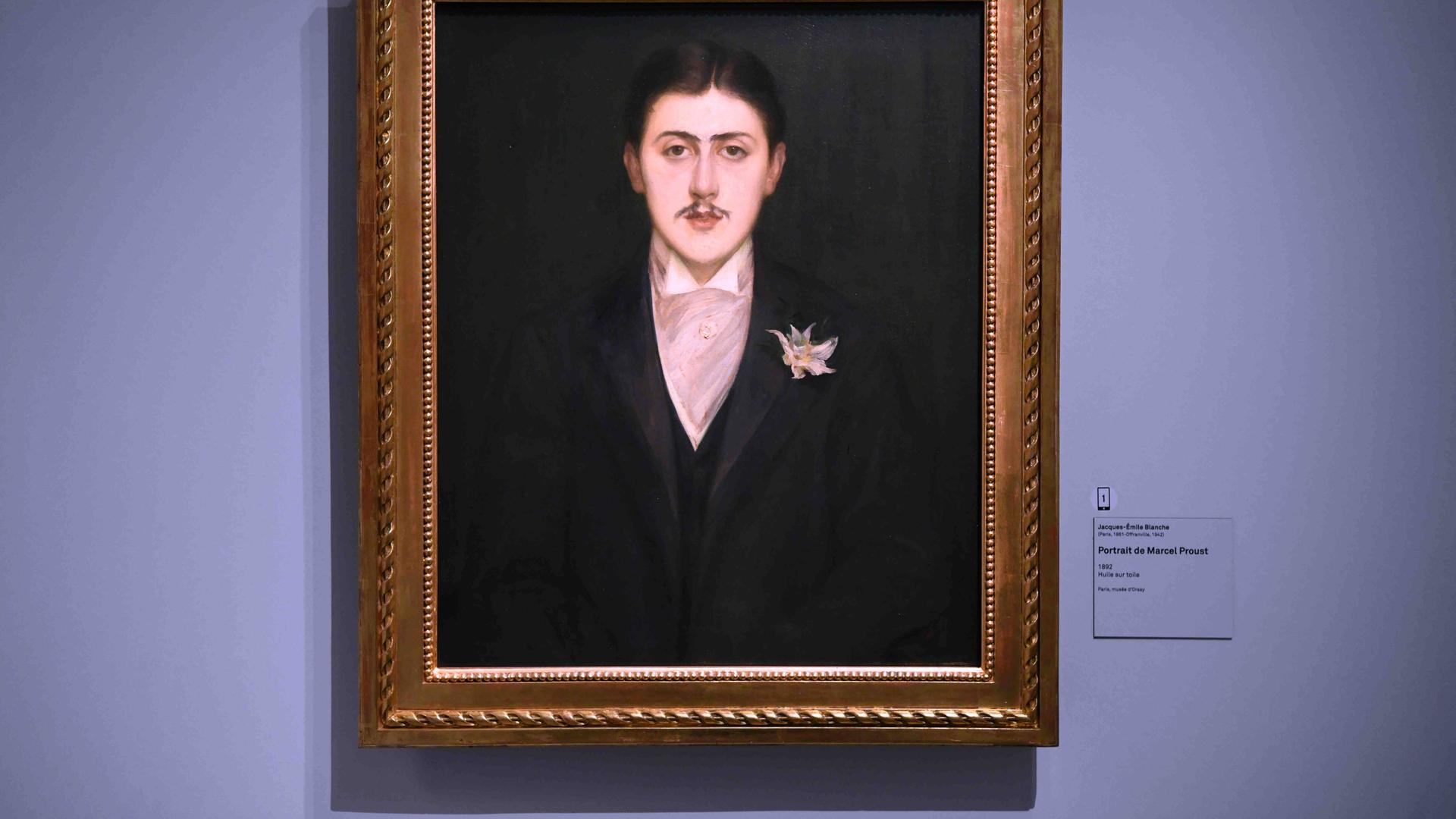 Ein gerahmtes Portraitbild des französischen Schriftstellers Marcel Proust hängt in einem Rahmen an einer violetten Wand. Daneben hängt eine Beschriftung des Bildes.