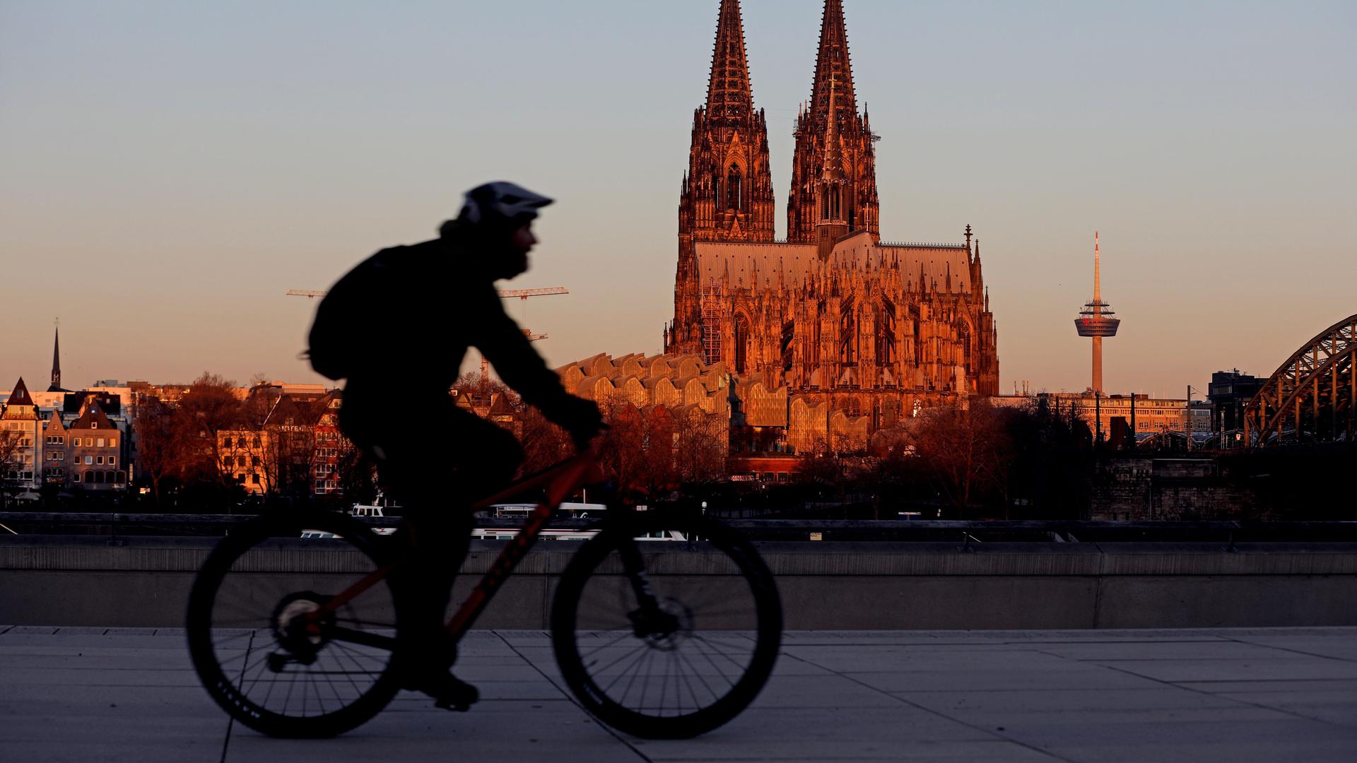 Ein Radfahrer fährt am Morgen am Kölner Dom vorbei, der von der aufgehenden Sonne angeleuchtet wird.