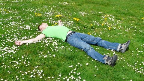 Ein Mann in Jeans und grünem Shirt liegt ausgestreckt mit dem Rücken auf einer blühenden Wiese und schläft.